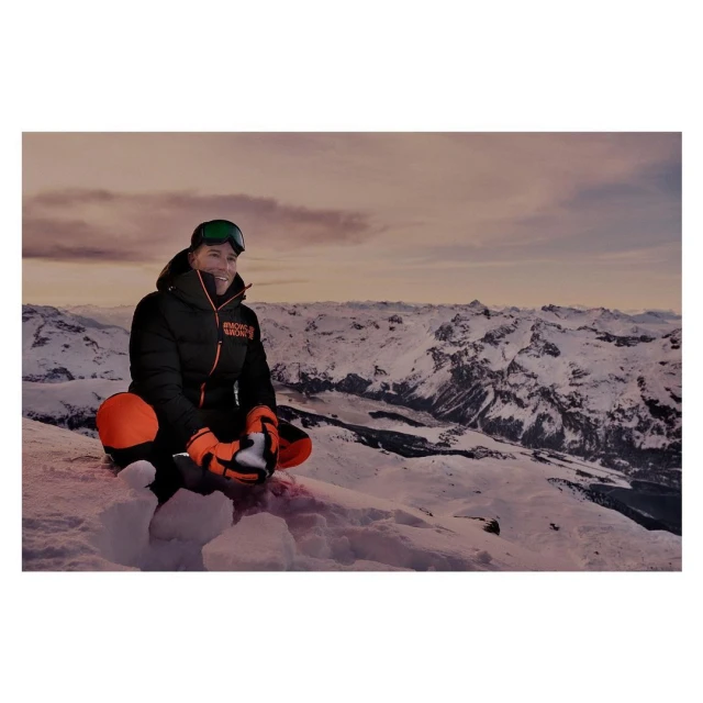Woman wearing ski goggles and winter apparel sits cross-legged in a snowy, mountainous outdoor environment. Scenic landscape in the distance.