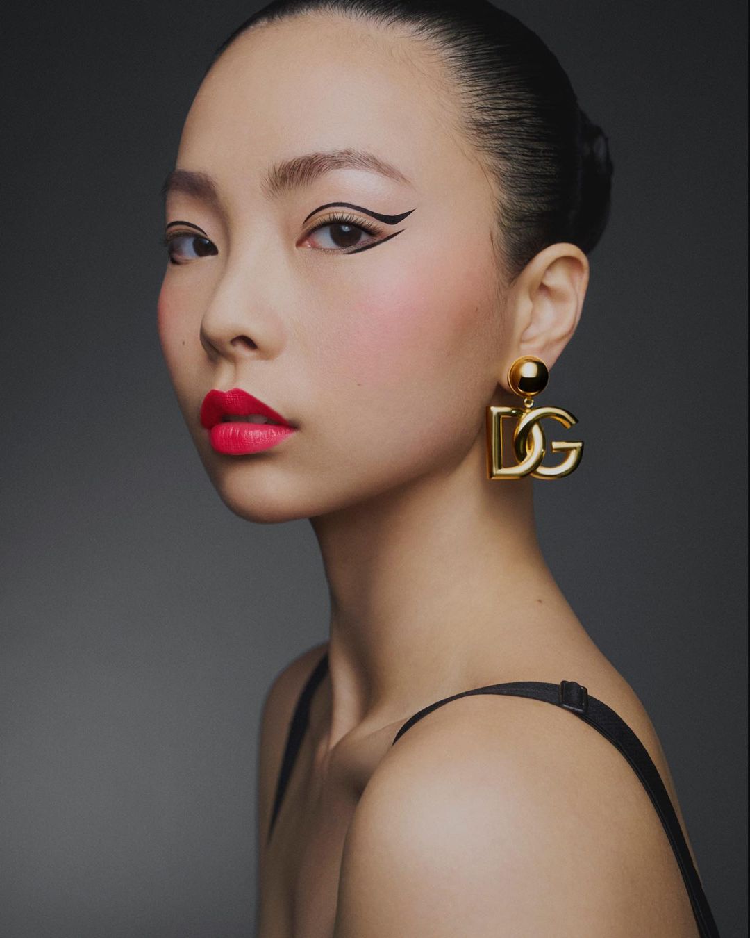 Close-up beauty shot of a young Asian woman with dramatic winged eyeliner and bright red lips wearing large gold Dolce & Gabbana earrings.