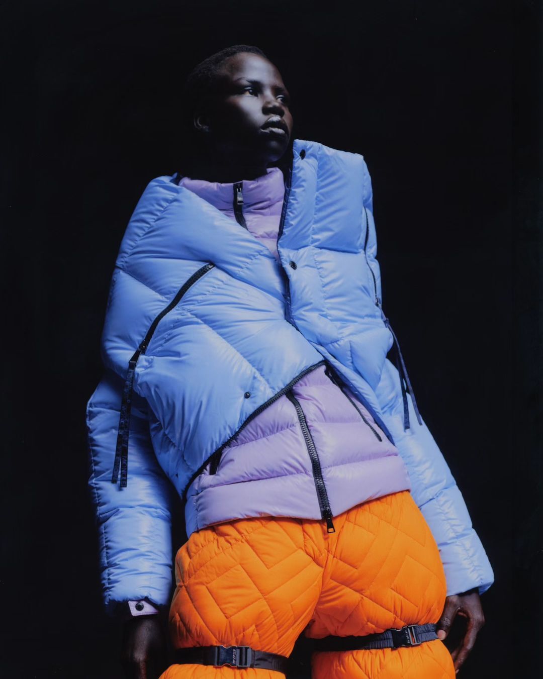Fashion shot of an African American adult wearing a light blue puffer jacket with orange quilted pants in a dark studio setting.