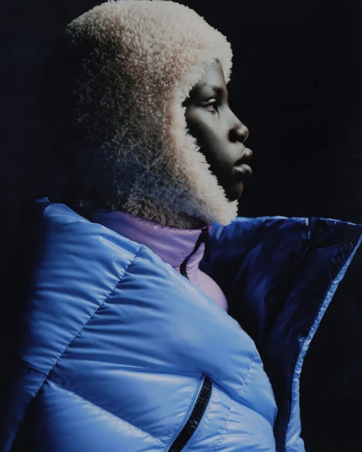 Profile of an African-descent adult in a light blue puffer jacket and fuzzy hat against a dark background, gazing into the distance.