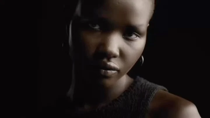 Close-up portrait of a Black woman with dark hair, dark eyes, and silver hoop earrings against a black background. The lighting is dramatic.