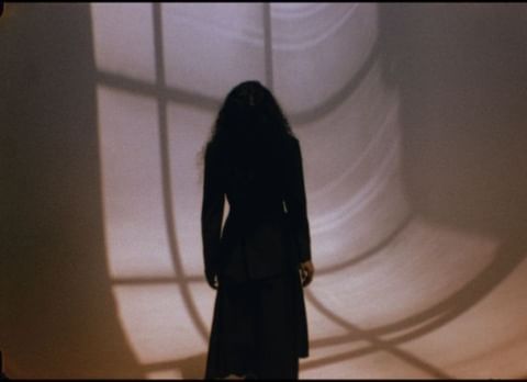 Adult female figure stands in shadow against a wall, highlighted by a patterned light design in shades of beige, brown, and black.