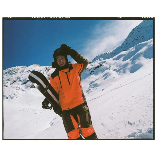 A snowboarder in an orange jacket stands on a snowy mountain slope under a blue sky, holding a snowboard and wearing a helmet.