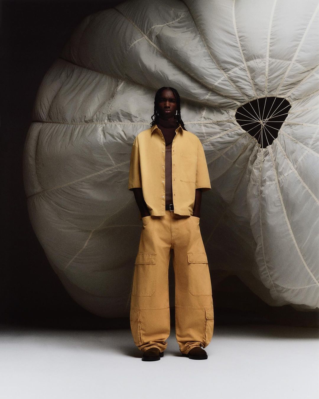 Male fashion model poses in tan shirt and wide-leg pants in front of parachute in studio for fashion editorial photoshoot.