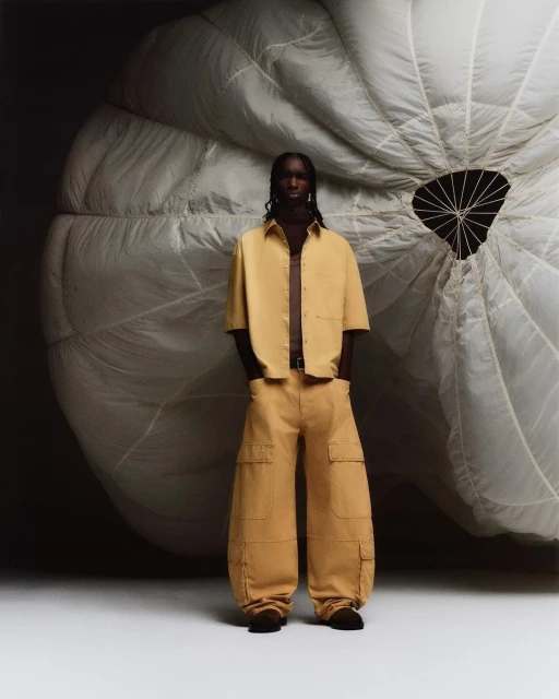 Male fashion model poses in tan shirt and wide-leg pants in front of parachute in studio for fashion editorial photoshoot.
