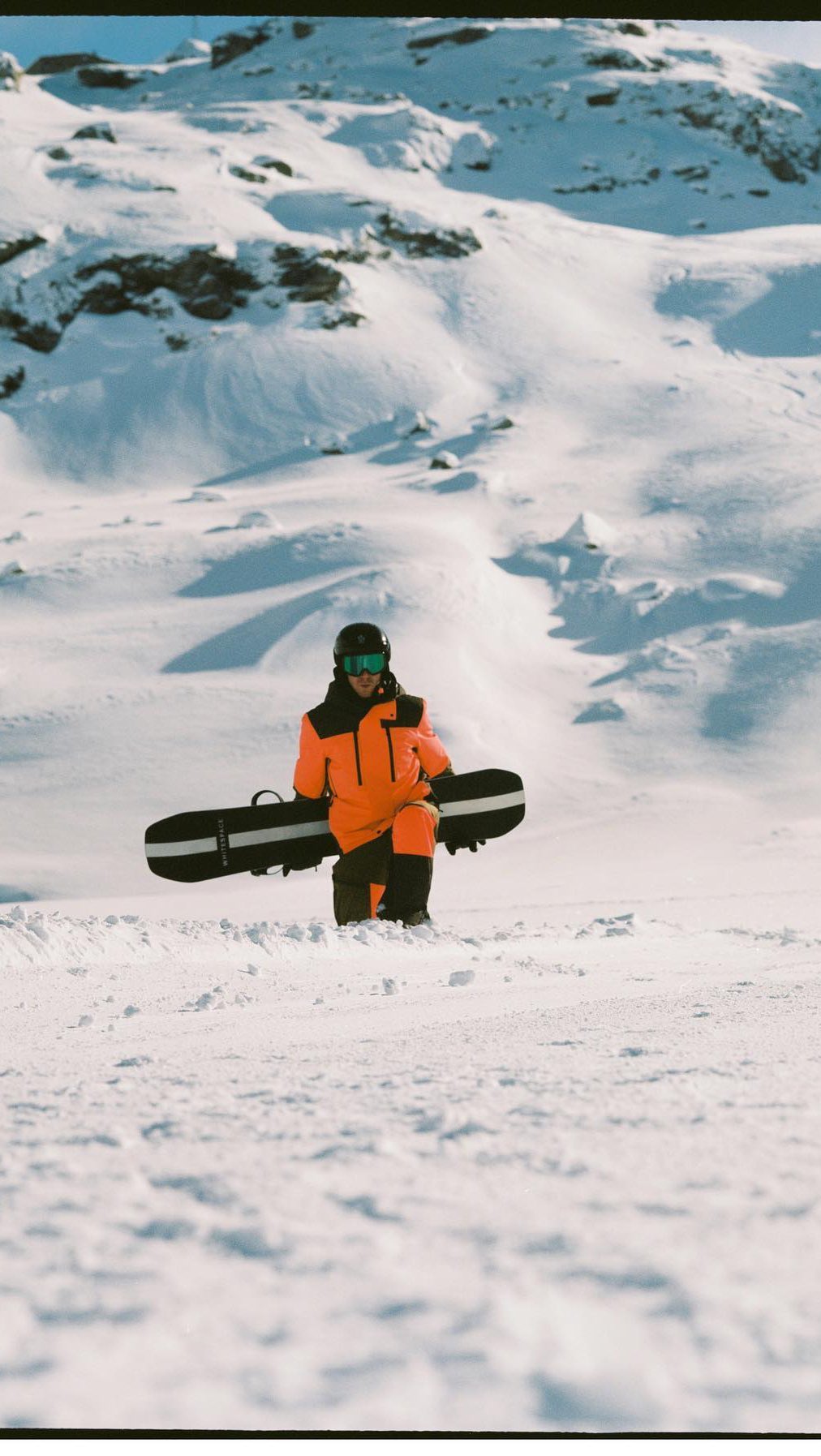 Person in bright orange snowsuit carries snowboard on snowy mountain landscape. Winter sports apparel for snowboarding.