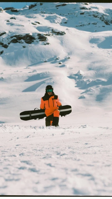 Person in bright orange snowsuit carries snowboard on snowy mountain landscape. Winter sports apparel for snowboarding.