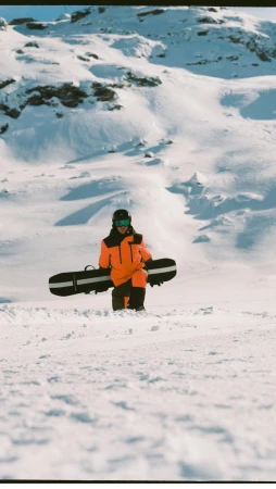 Person in bright orange snowsuit carries snowboard on snowy mountain landscape. Winter sports apparel for snowboarding.