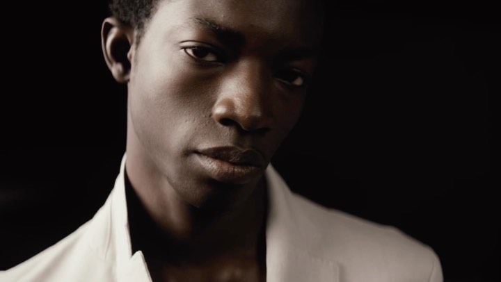 Portrait of a young Black man wearing a white blazer against a black backdrop, showcasing fashion and beauty in a studio setting.