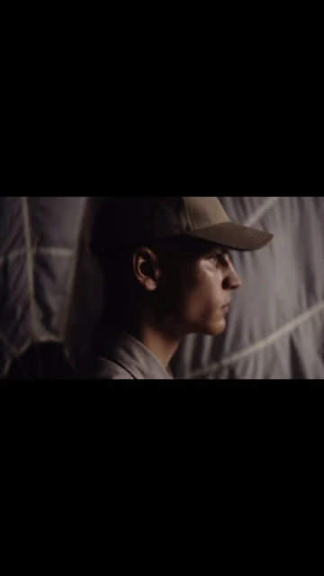 Profile of a man wearing a cap, with neutral expression, standing indoors against a textured background in a low-light, muted setting.