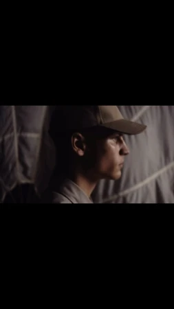 Profile of a man wearing a cap, with neutral expression, standing indoors against a textured background in a low-light, muted setting.