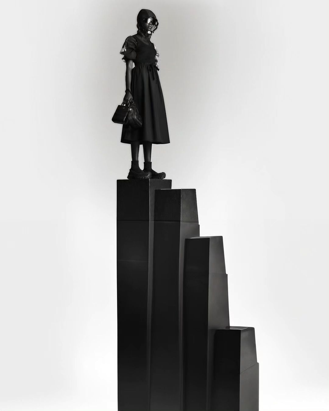 Woman in dark dress and gas mask poses atop black geometric steps in a studio setting, creating a surreal, monochromatic fashion image.