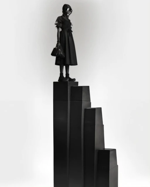 Woman in dark dress and gas mask poses atop black geometric steps in a studio setting, creating a surreal, monochromatic fashion image.