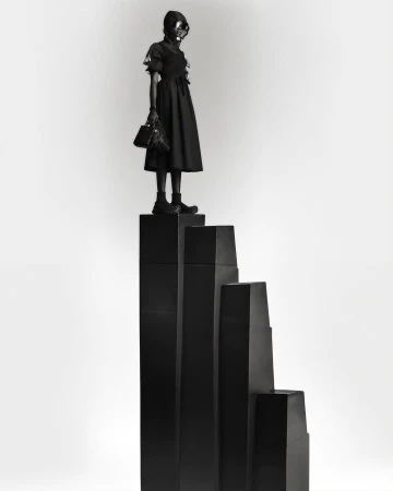 Woman in dark dress and gas mask poses atop black geometric steps in a studio setting, creating a surreal, monochromatic fashion image.