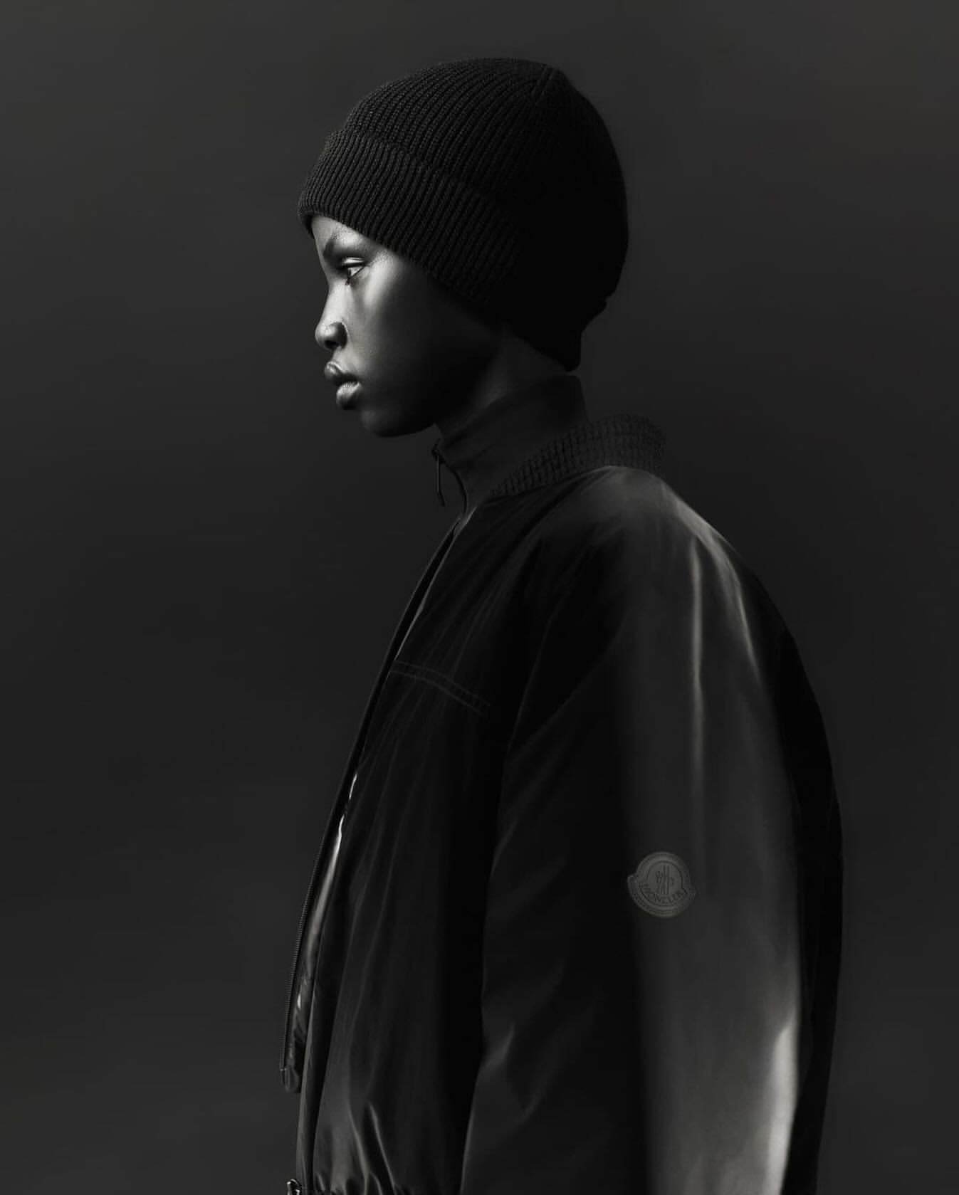 Profile of a young Black adult wearing a black beanie and a stylish black jacket, showcasing a fashion-forward look against a dark backdrop.