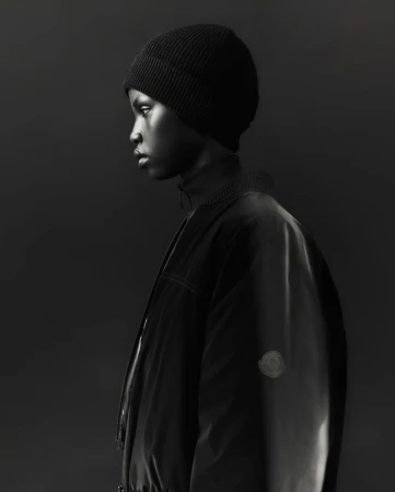 Profile of a young Black adult wearing a black beanie and a stylish black jacket, showcasing a fashion-forward look against a dark backdrop.