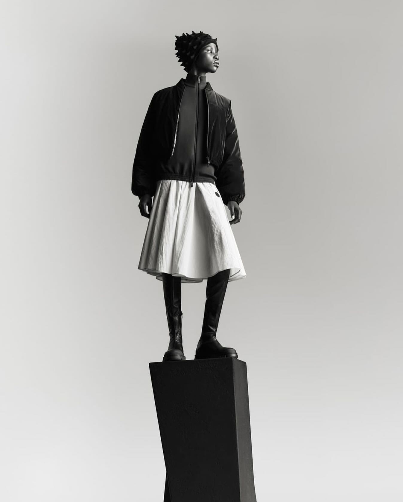 Full-length shot of an adult woman in a black jacket, white skirt, and boots, standing on a black geometric pedestal against a grey backdrop.