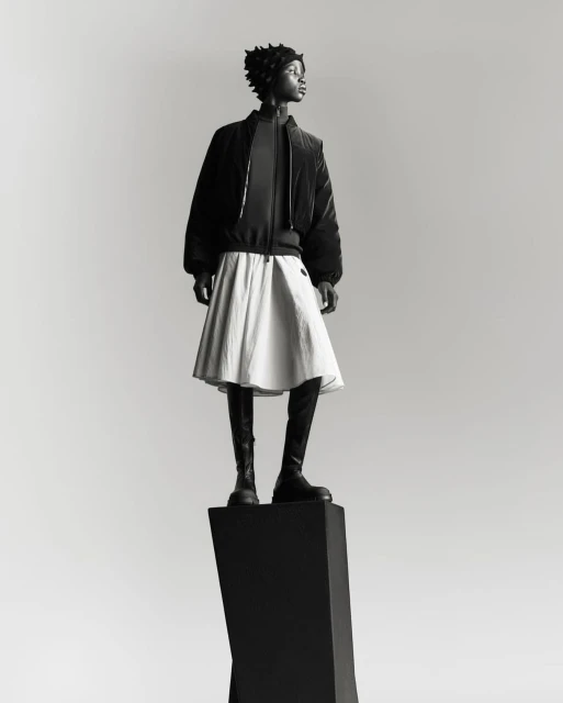 Full-length shot of an adult woman in a black jacket, white skirt, and boots, standing on a black geometric pedestal against a grey backdrop.