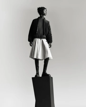 Full-length shot of an adult woman in a black jacket, white skirt, and boots, standing on a black geometric pedestal against a grey backdrop.
