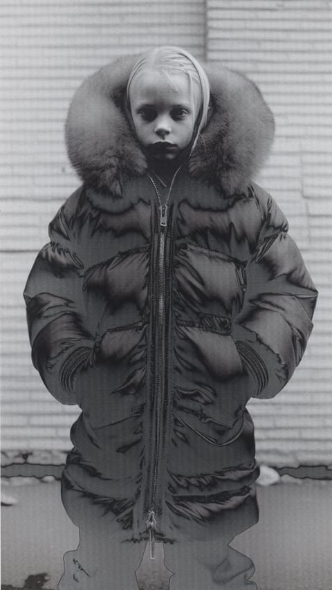 Child in a puffy coat with a fur-trimmed hood, portrait style. The subject stands against a wall of light and dark grey tones.