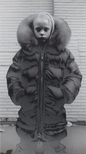 Child in a puffy coat with a fur-trimmed hood, portrait style. The subject stands against a wall of light and dark grey tones.