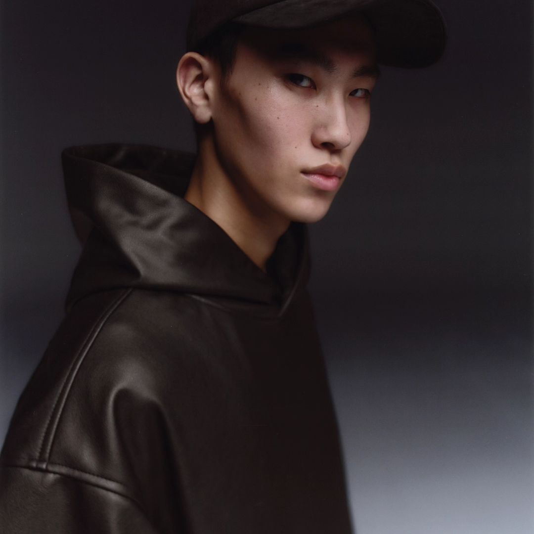 Portrait of a young adult wearing a brown hooded leather-look top and baseball cap against a dark grey studio backdrop.