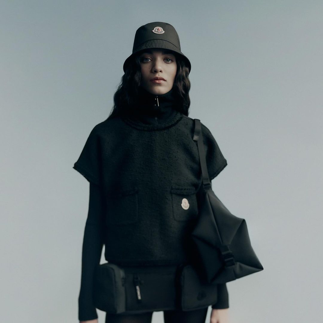 Woman wearing black bucket hat and matching outfit with black bag, posing against a pale blue background in a studio setting.