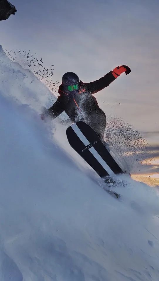 A snowboarder carving through fresh powder on a sunny mountain slope, wearing a black jacket, helmet, and bright orange gloves.