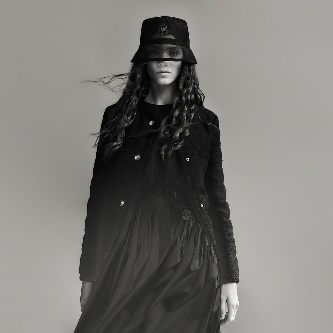 Monochrome studio shot of a fashionable adult woman in a bucket hat and black dress, with a shearling jacket and long, wavy hair.