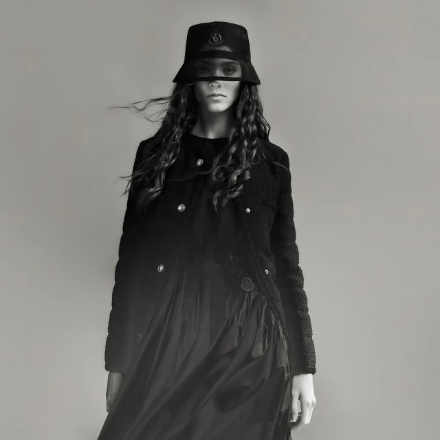 Monochrome studio shot of a fashionable adult woman in a bucket hat and black dress, with a shearling jacket and long, wavy hair.