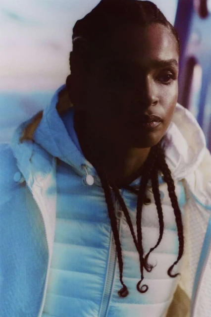 Close-up portrait of a woman with dark hair in braids wearing a hooded down jacket, with soft lighting and a blurred background.