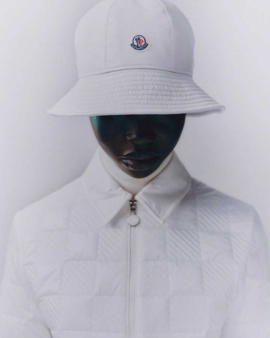 Person wearing a Moncler bucket hat and a quilted jacket in a studio setting, showcasing a minimalist fashion aesthetic.