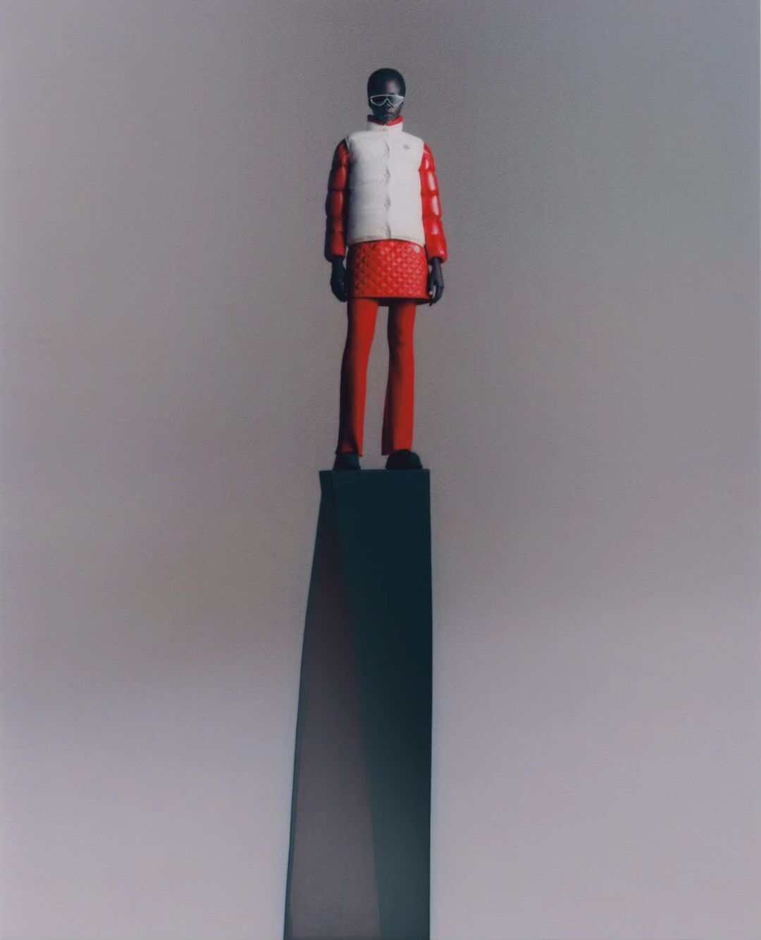 Full shot of person standing on black sculpture against grey backdrop, wearing goggles, white vest, red top, and red pants.
