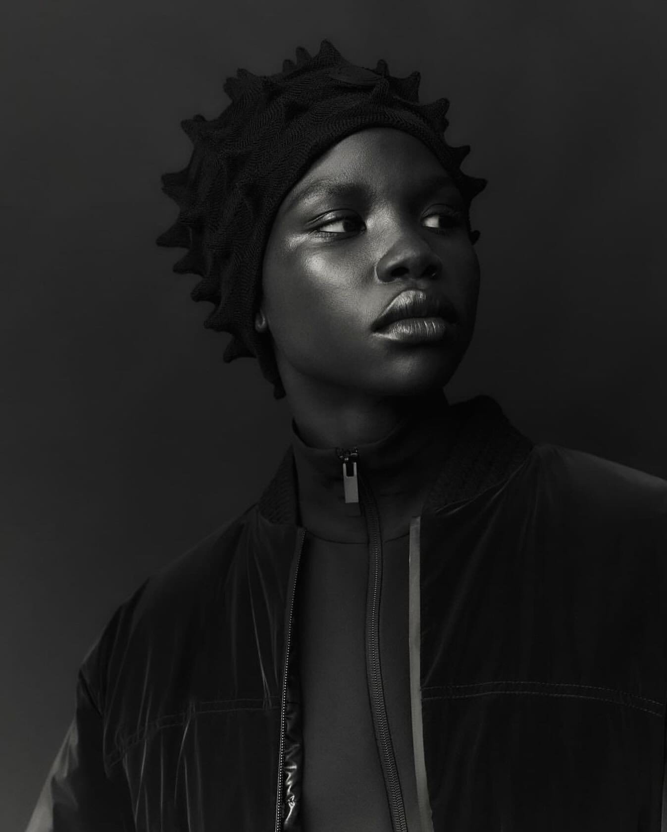 Close-up portrait of an African American woman wearing a black, textured hat and jacket, set against a dark monochrome background.