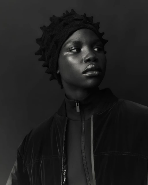 Close-up portrait of an African American woman wearing a black, textured hat and jacket, set against a dark monochrome background.