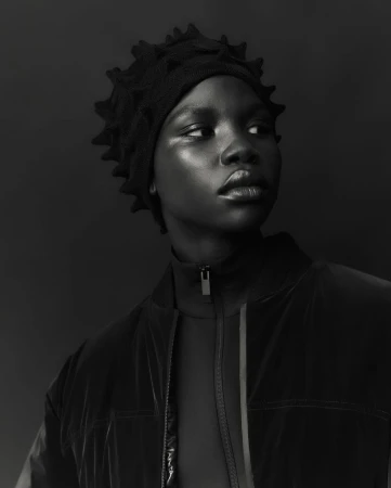 Close-up portrait of an African American woman wearing a black, textured hat and jacket, set against a dark monochrome background.