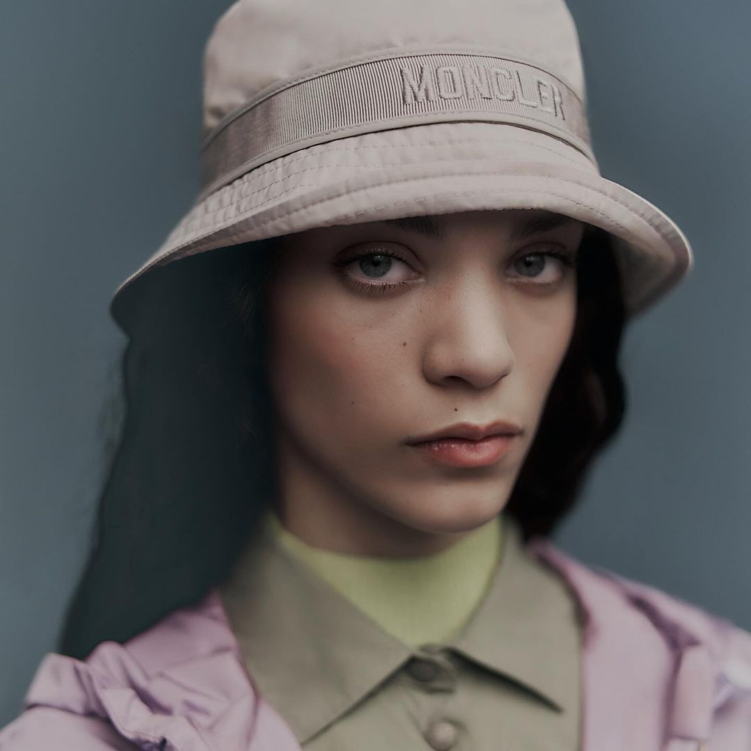 Close-up portrait of a woman wearing a Moncler bucket hat and a buttoned shirt, with soft lighting and a neutral-toned background.