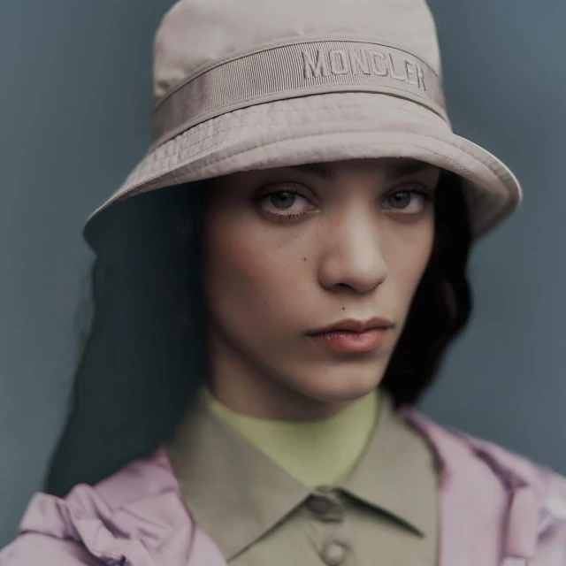 Close-up of a woman wearing a beige Moncler bucket hat with a pale green shirt and lilac jacket against a muted blue background.