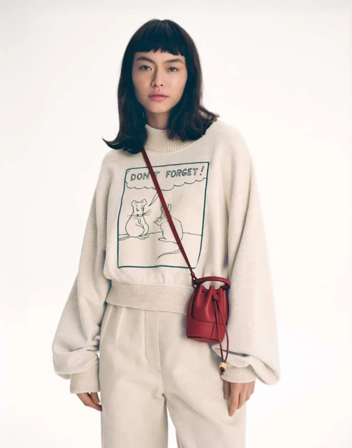 Asian woman in a studio wearing a sweatshirt with a cartoon graphic, paired with cream trousers and a small red leather bucket bag.