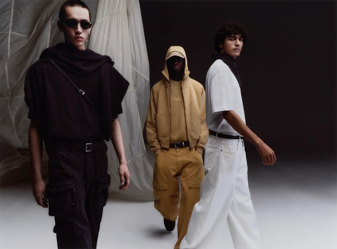 Three adult male fashion models present apparel in a studio setting with dark and neutral backgrounds. Light shines evenly on them.
