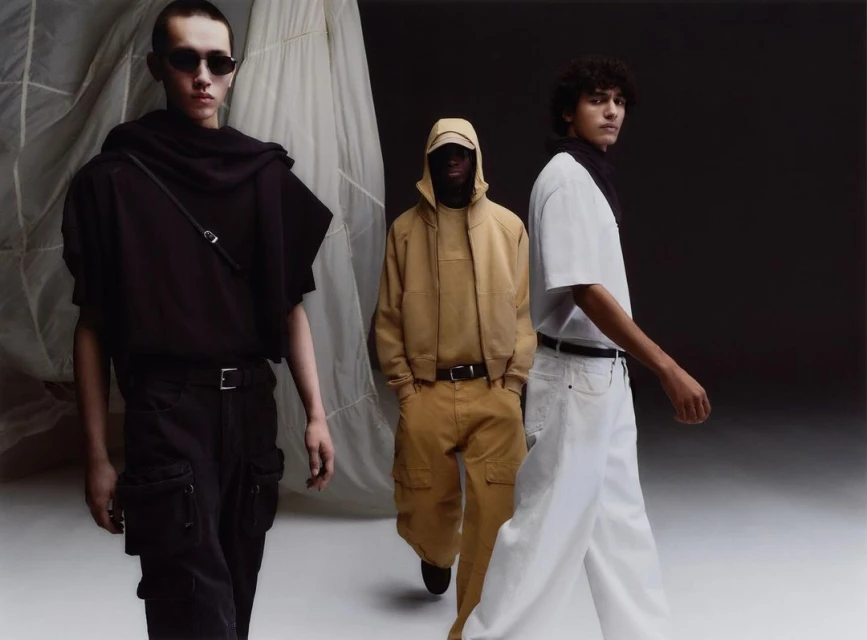 Three adult male fashion models present apparel in a studio setting with dark and neutral backgrounds. Light shines evenly on them.