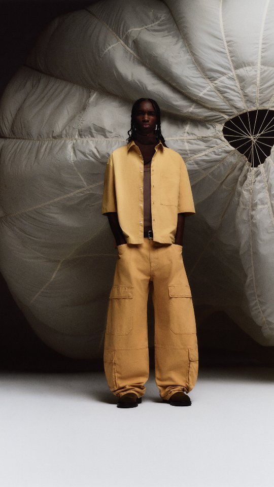 A man in a golden yellow shirt and baggy cargo pants stands in front of an inflated parachute in a fashion editorial shot.