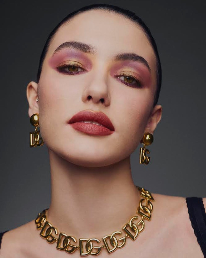Close-up portrait of a young adult woman with pink and gold eyeshadow, red lipstick, and gold Dolce & Gabbana jewelry against a gray background.