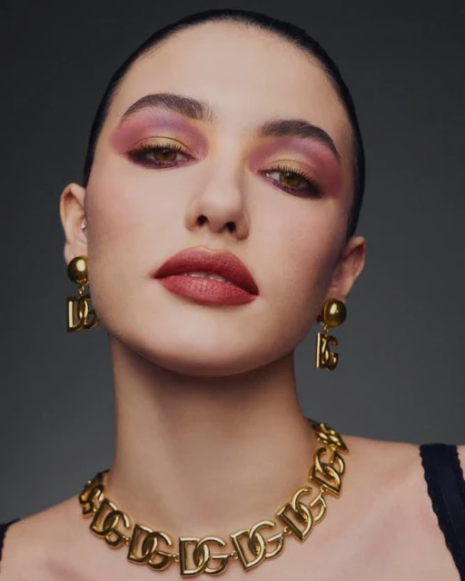 Close-up portrait of a young adult woman with pink and gold eyeshadow, red lipstick, and gold Dolce & Gabbana jewelry against a gray background.