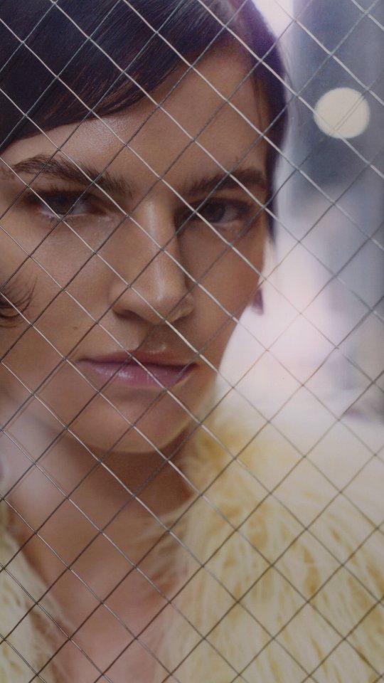 Close-up portrait of a woman with short dark hair and light skin, wearing a textured yellow jacket, posing behind a wire mesh.