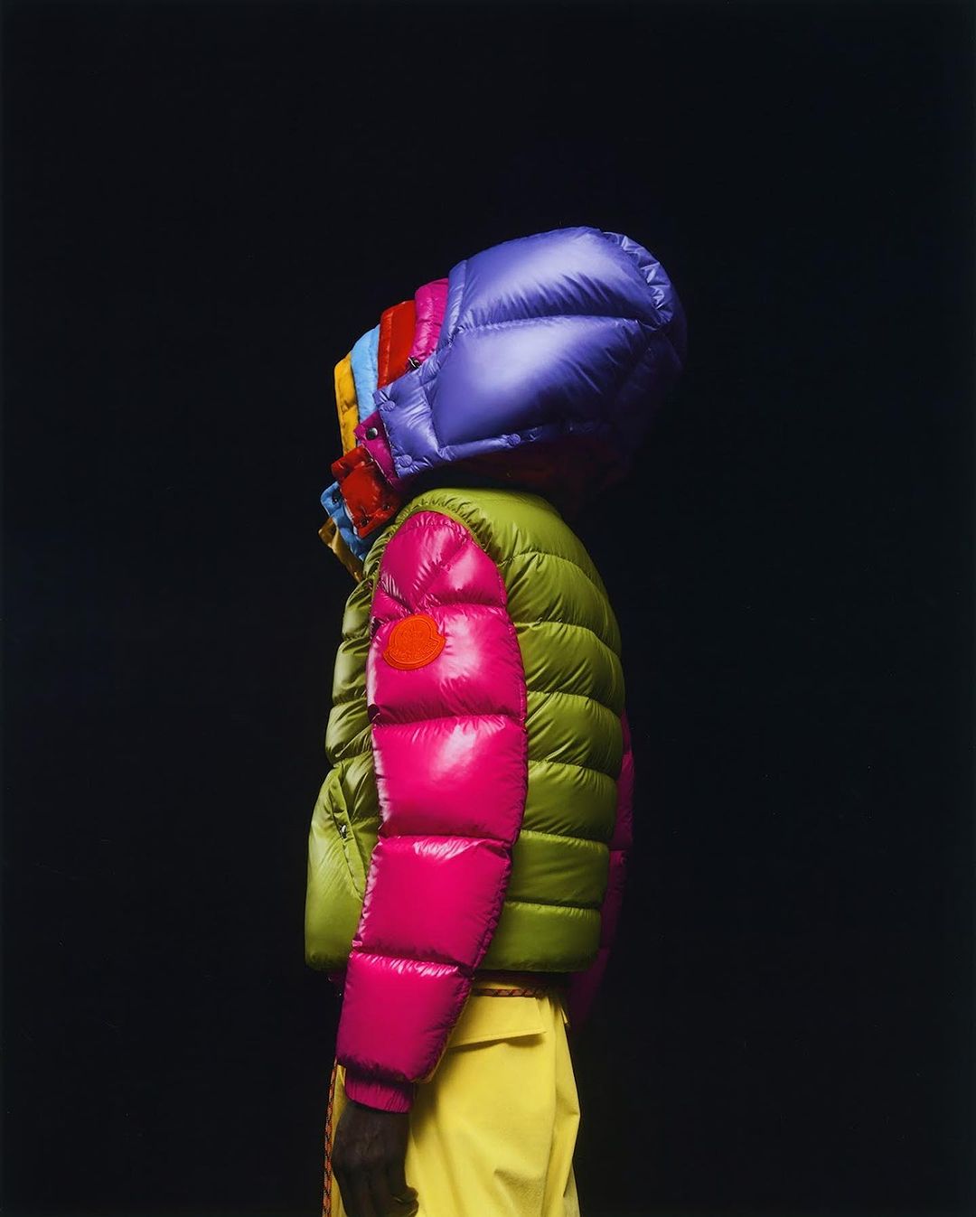 A person wearing a puffer jacket with a colorful hood, shot in a photo studio against a black backdrop. The jacket features bright colors.