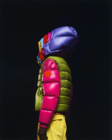 A person wearing a puffer jacket with a colorful hood, shot in a photo studio against a black backdrop. The jacket features bright colors.