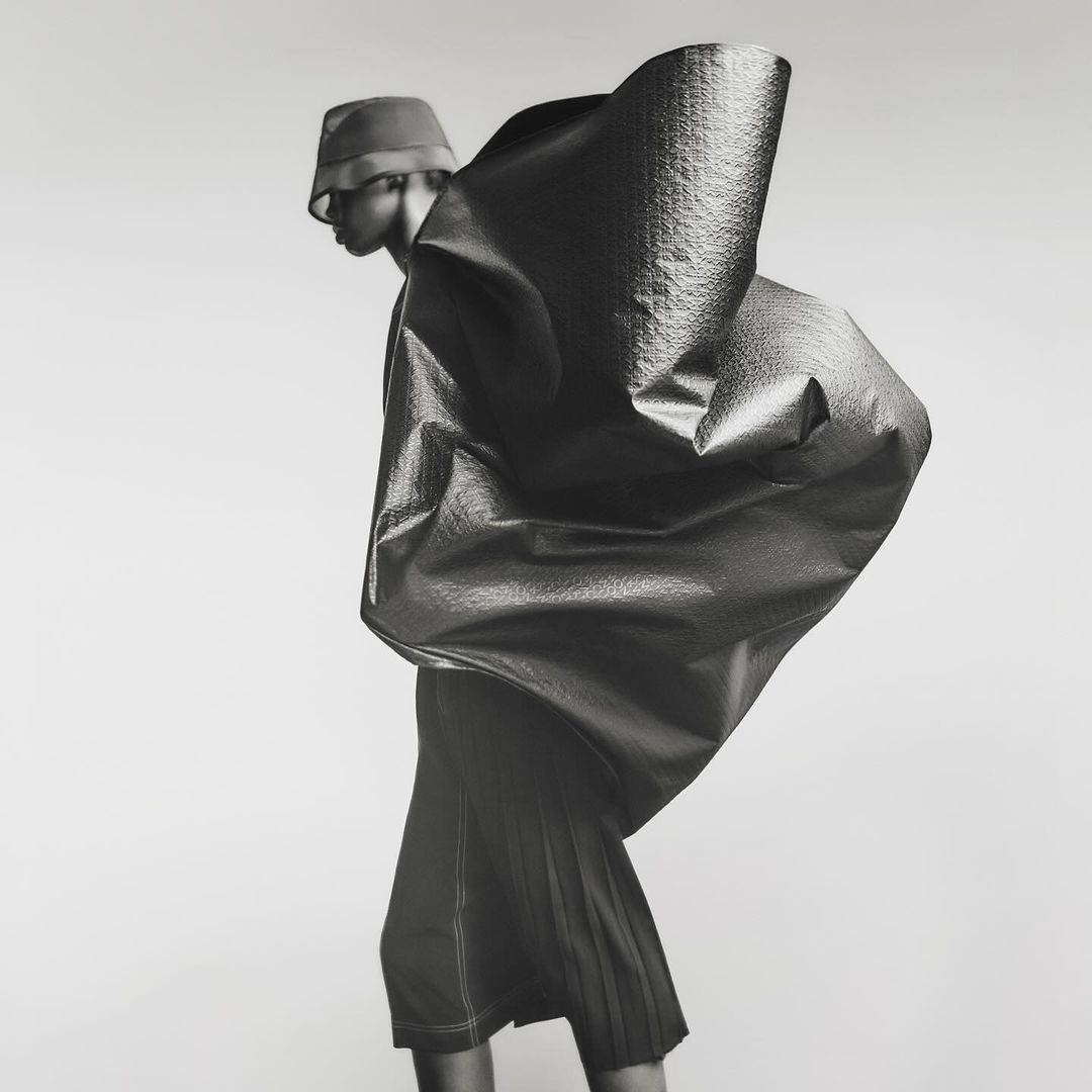 Fashion photo of a person wearing a bucket hat and a large, sculptural garment with a textured surface, set against a neutral background.