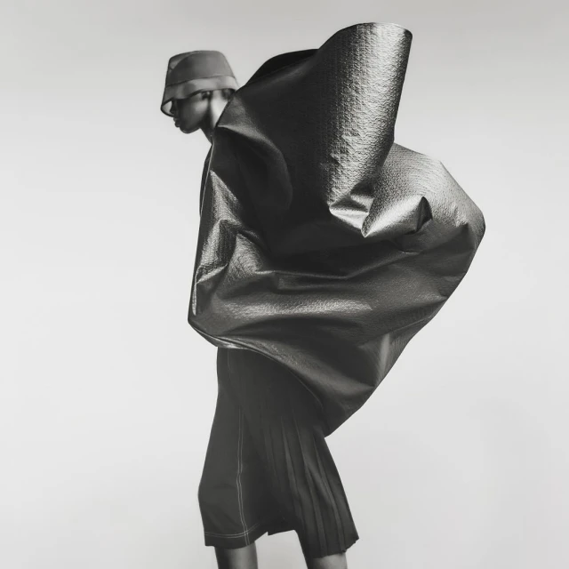 Fashion photo of a person wearing a bucket hat and a large, sculptural garment with a textured surface, set against a neutral background.