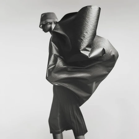 Fashion photo of a person wearing a bucket hat and a large, sculptural garment with a textured surface, set against a neutral background.